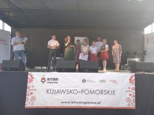 Oficjele na scenie wręczaja nagrodę grupie kobiet ubranych w stroje ludowe